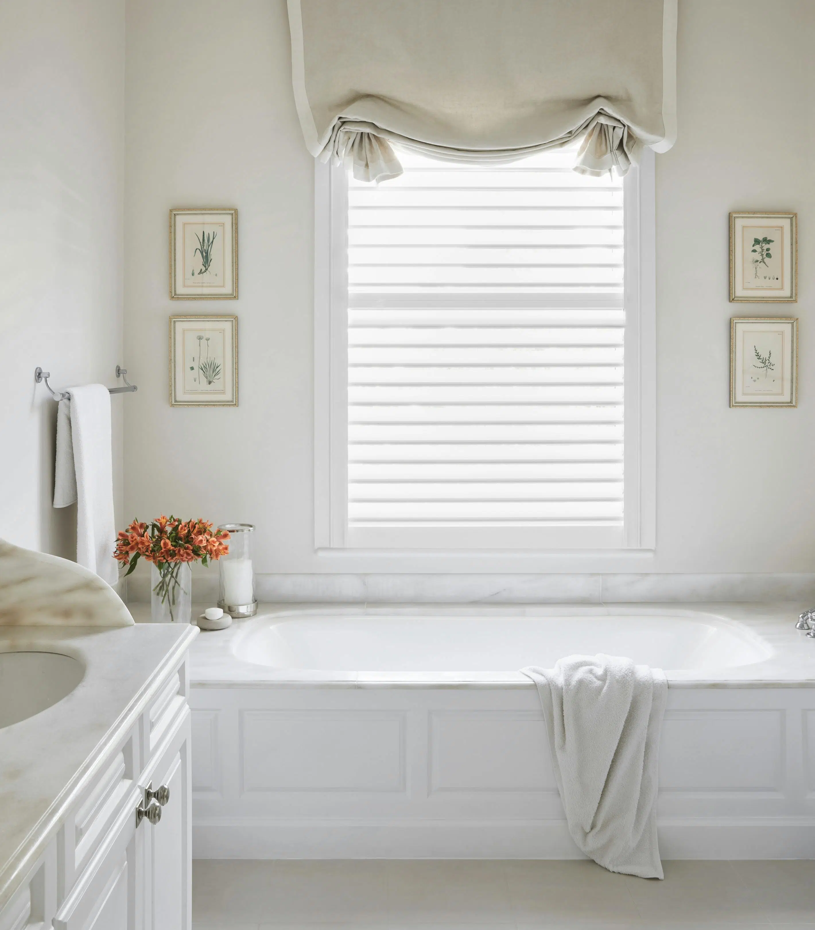 A bathroom in soft, white tones with a bathtub in the centre, frames on the wall and a basin to the left.