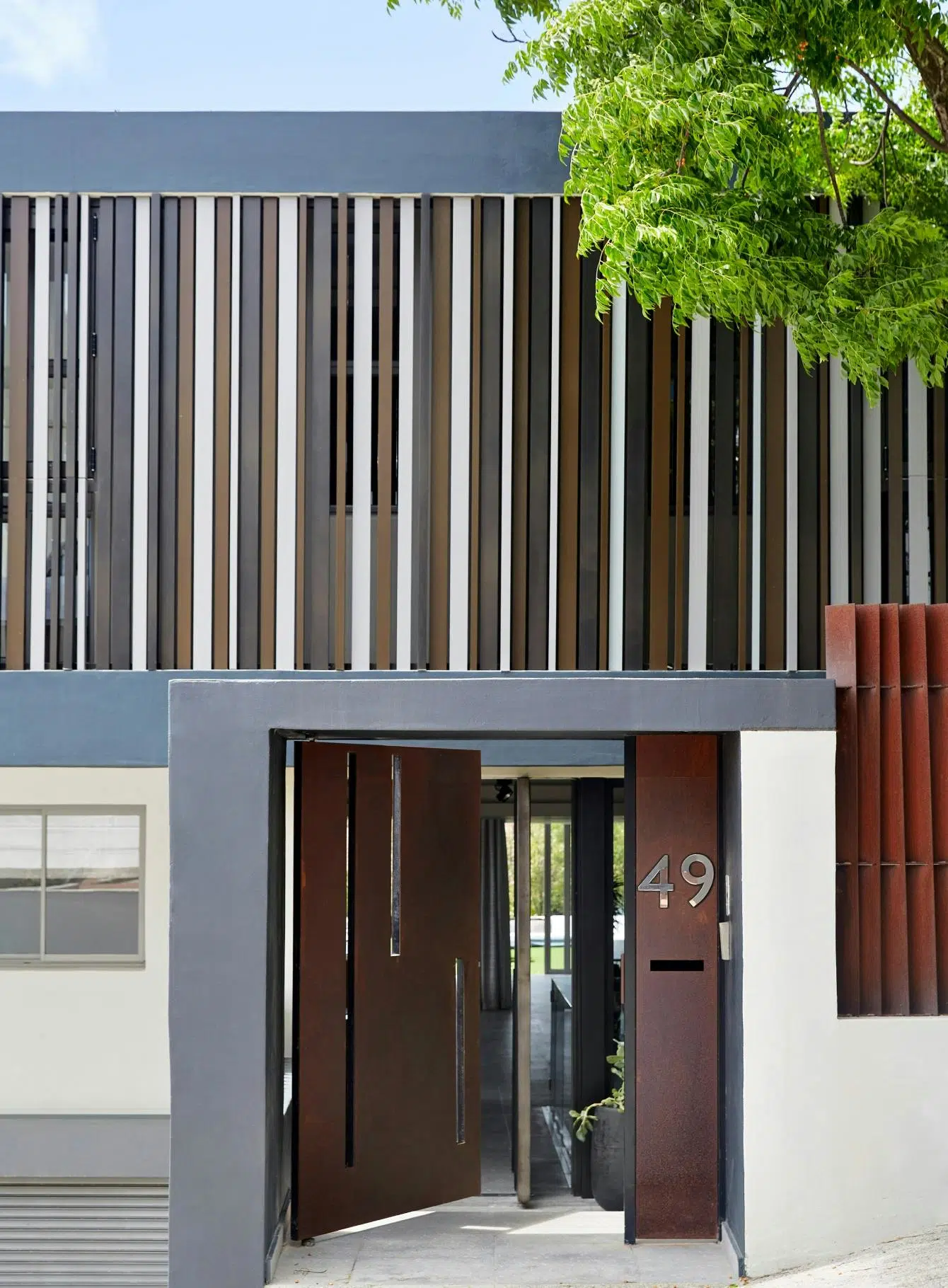 The exterior of a home that features a panelled façade in brown, earthy tones and white, with an ajar front door leading into the house.